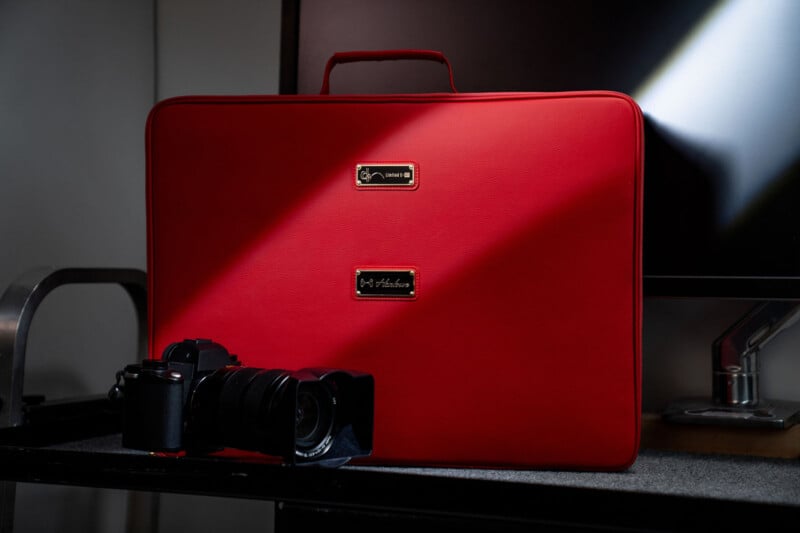 A red leather briefcase with a handle stands upright on a surface, with a black camera placed in front of it. Sunlight creates a diagonal highlight across the case. A monitor is partially visible in the background.