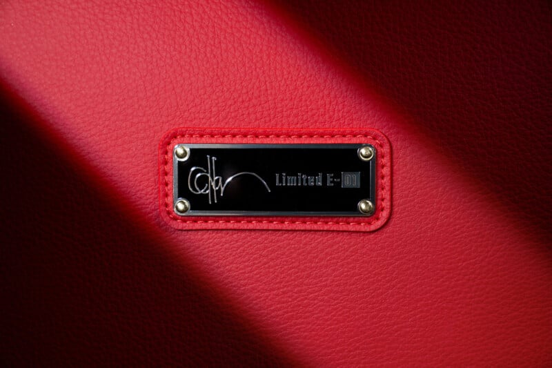 A close-up of a black and silver metal label on red textured leather. The label reads "Limited E-01" and has a signature-like design, attached with four gold screws.