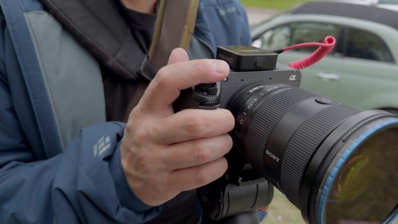A person wearing a blue jacket holds a Sony Alpha camera with both hands, preparing to take a photo. A car and greenery are visible in the blurred background.