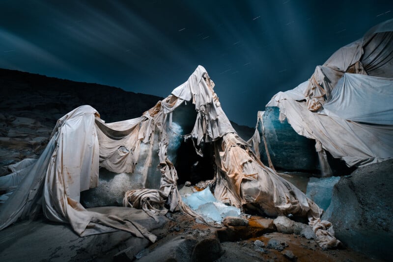 Large sheets of fabric drape over melting ice and rocks under a night sky with streaking clouds and faint star trails, creating a surreal and haunting landscape.