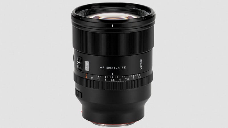 A black camera lens with a red ring near the mount, labeled "AF 85/1.4 FE," featuring focus and aperture markings, and various switches on the side, set against a plain light gray background.