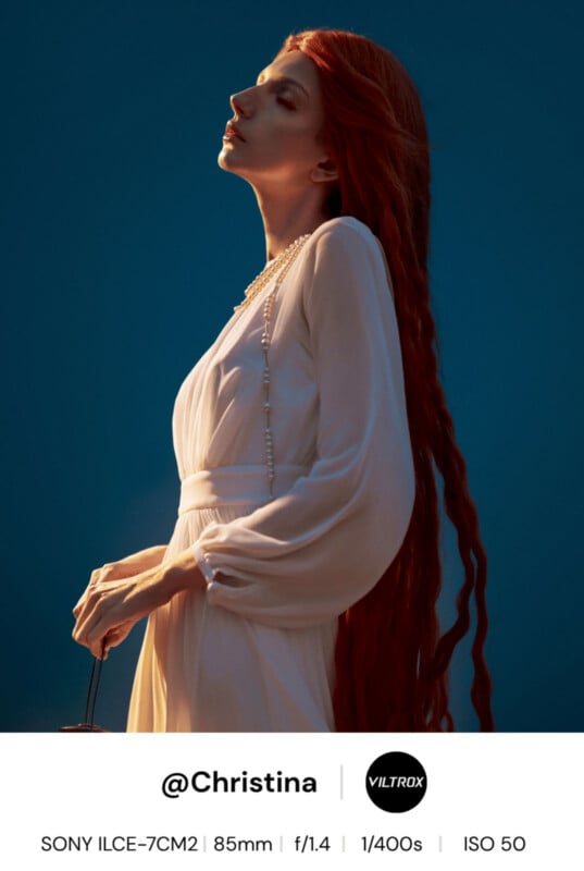 A woman with long red hair and a white flowing dress stands in profile against a deep blue background, softly lit. She holds a pearl necklace and looks upward with a serene expression. Camera and social info are overlaid below.