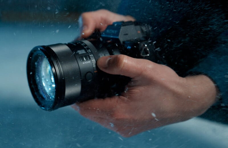 A close-up of a person’s hands holding a camera with water droplets splashing around, suggesting the camera is being used in wet conditions.