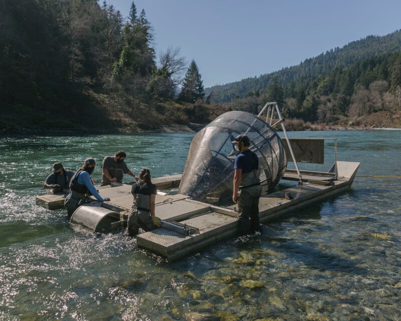 Five people work around a floating fish trap structure in a clear river, surrounded by forested hills under a clear blue sky. The group appears to be conducting river monitoring or research activities.