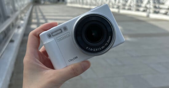 A hand holding a white digital camera outdoors. The camera has the brand name "VASTfca" and model "City100" printed on it. The lens details include "F1.6-2.8/3.5-10.35mm." A blurred walkway is visible in the background.