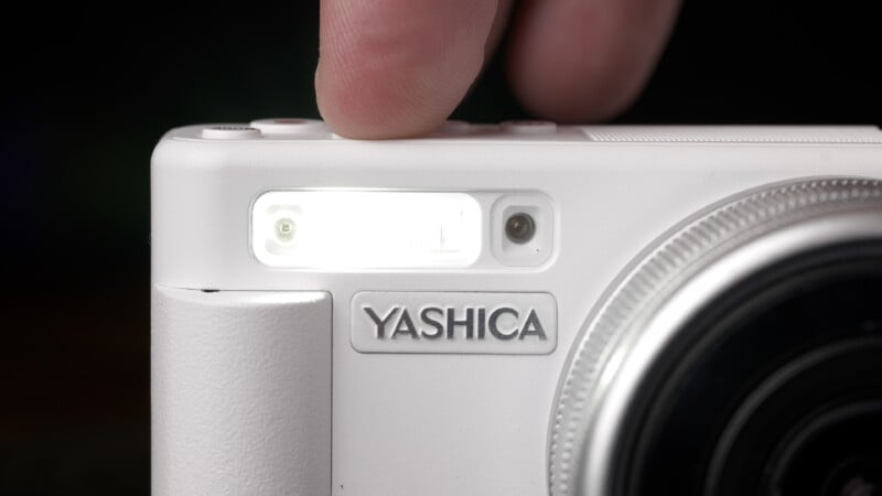 A close-up of a white Yashica camera with a finger pressing a button, activating the camera’s flash. The Yashica logo is clearly visible above the grip.