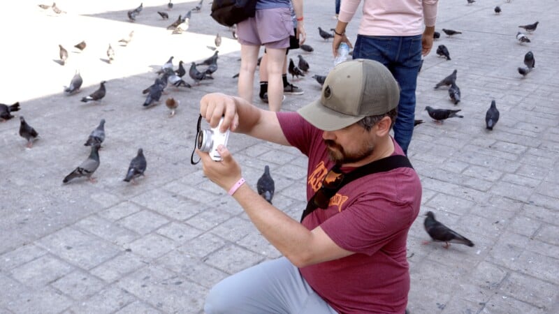 A man in a red shirt and cap kneels on a stone street, taking a photo with a camera as pigeons walk and fly around him. Other people and pigeons are visible in the background.