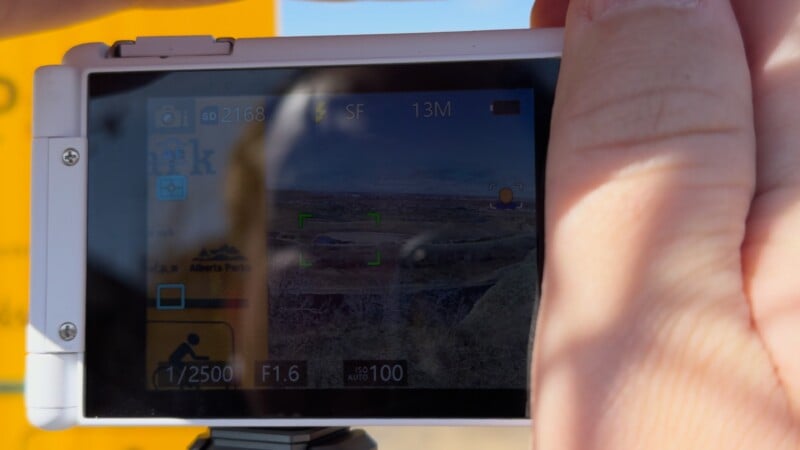 Close-up of a person’s hands holding a digital camera, with the camera’s screen displaying settings and a landscape view. The background is slightly blurred and shows part of a yellow sign.