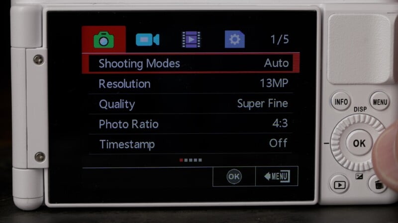 Close-up of a digital camera screen showing settings menu. "Shooting Modes" is set to Auto, resolution to 13MP, quality to Super Fine, photo ratio to 4:3, and timestamp is off. A finger is near the control wheel.